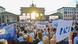 A crowd of people by the Brandenburg Gate holding up banners reading "peace" in different languages, a flag with a dove and the word "Egidio" A crowd of people by the Brandenburg Gate holding up banners reading "peace" in different languages, a flag with a dove and the word "Egidio"