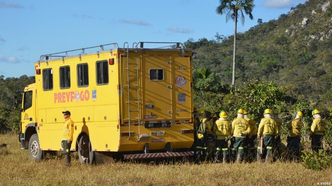 Caminhão amarelo e pessoas com roupas amarelas equipadas para combater incêndio em uma área de mata