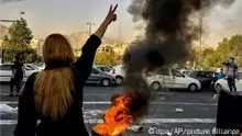 30.9.2022, Eine Frau steht während einer Demonstration nach dem Tod der 22-jährigen Mahsa Amini vor einem brennenden Autoreifen und zeigt das Victory-Zeichen. Am 16. September jährt sich erstmals der Tod der Kurdin Jina Mahsa Amini, der im vergangenen Jahr im Iran die schwersten Proteste seit Jahrzehnten auslöste. Die Staatsführung tritt wieder selbstbewusst auf. Doch die Gesellschaft im Land steuert auf ungewisse Zeiten zu. (zu dpa «Ein Jahr nach den Protesten: Iran zwischen Wandel und Stillstand»)