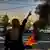 Një grua me shpinë nga kamera, dorën e djathtë të e ka të ngritur dhe me dy gishtat ka bërë shenjën V. Ajo është në buzë të rrugës dhe para saj sapo ka shpërthyer një bombë “Molotov”. 
