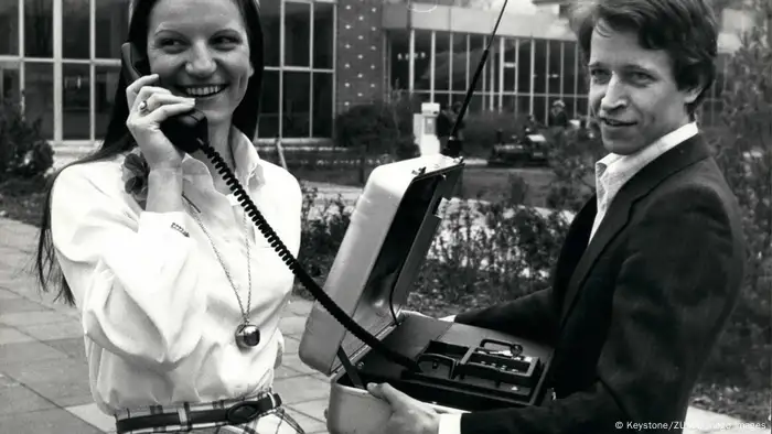 Eine Frau hält einen Telefonhörer ans Ohr, der über ein Kabel mit einem Koffer verbunden ist, den ein Mann in beiden Händen hält.