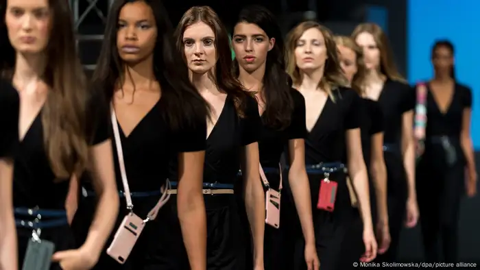 Eine Reihe von Frauen in schwarzen Kleidern läuft bei einer Fashion Show mit über die Schultern gehängten Mobiltelefonen über den Laufsteg.