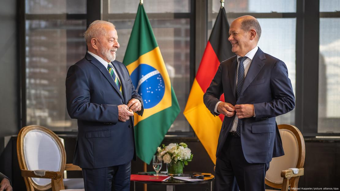 Lula e Scholz abotam os paletós e sorriem em frente a uma bandeira do Brasil e uma da Alemanha