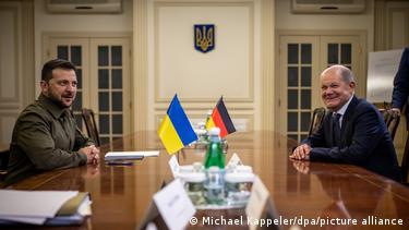 Η εικόνα δείχνει τον Ουκρανό Πρόεδρο Ζελένσκι να κάθεται στα αριστερά σε ένα σκούρο καφέ ξύλινο τραπέζι σε μια συνάντηση με τον Γερμανό Καγκελάριο Σολτς στη Νέα Υόρκη μετά από μια συνεδρίαση του Συμβουλίου Ασφαλείας του ΟΗΕ το 2023. Στο τραπέζι υπάρχει μια ουκρανική και μια γερμανική σημαία