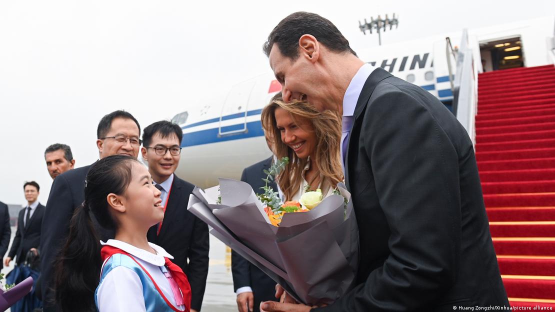 Presidente da Síria, Bashar al-Assad, e esposa são recebidos por uma menina chinesa no aeroporto de Hangzhou. A escada do avião e a própria aeronave podem ser vistos ao fundo. Todos sorriem, e o presidente sírio segura um buquê de flores.