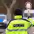 Polizist hält eine "Halt-Polizei-Kelle" nach oben, im Hintergrund ein nahendes Auto, daneben steht ein Polizei-Wagen