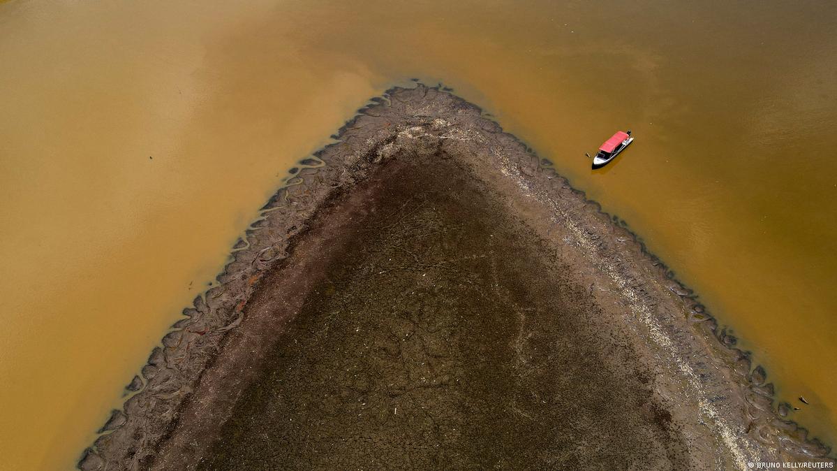 A triangular, dark brown headland juts out into the yellowish murky water; a red boat can be seen at the upper right edge of the image.