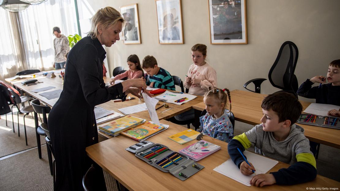 Eine blonde Lehrerin steht vor den Schulpulten mehrerer Kinder.