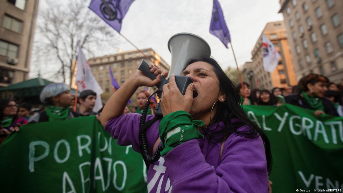 mujer habla con altavoz en manifestación callejera junto con otras mujeres, se ven banderas por el aborto legal.