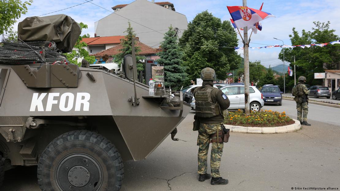 KFOR-i si garantues i paqes - ushtar i KFOR-it me kurriz nga kamera pranë një tanku, në sfond rruga me flamuj serbë dhe ushtarë të tjerë të KFOR