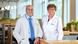 Nobel Prize winners Drew Weissman (left) and Katalin Kariko stand side by side in an office. Both wear white lab coats and smile at the camera.