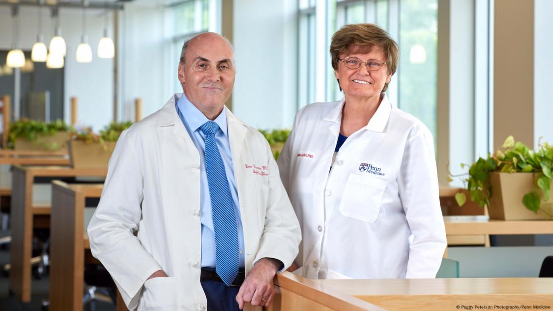 Nobel Prize winners Drew Weissman (left) and Katalin Kariko stand side by side in an office. Both wear white lab coats and smile at the camera.