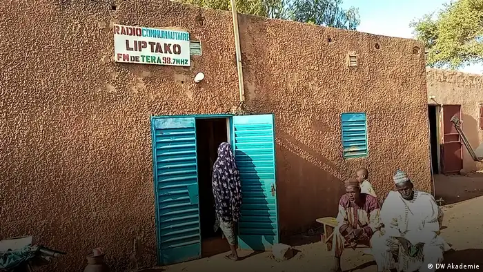 Photo shows several people sitting outside a building made of rammed-earth, with a bright blue door and a sign saying Liptako FM de Tera 98.7