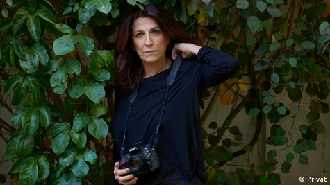 Mujer con cámara de fotos en la mano, vestida de negro, frente a pared con plantas de hojas verdes.