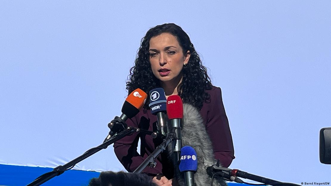 Presidentja e Kosovës Vjosa Osmani para mikrofonëve dhe një sfondi blu