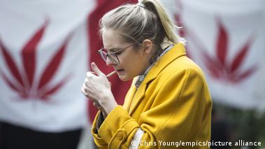 A blonde woman in a yellow coat lights up a joint in front of two Canadian flags where the maple leaf has been replaced with a cannabis leaf