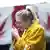 A blonde woman in a yellow coat lights up a joint in front of two Canadian flags where the maple leaf has been replaced with a cannabis leaf