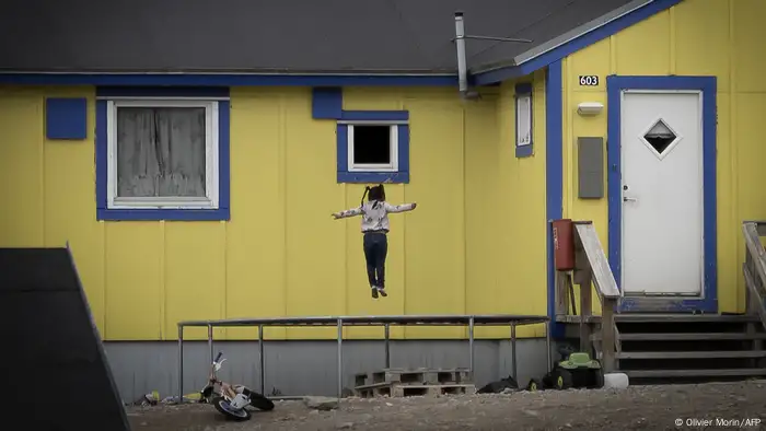 A girl bounces on a trampoline in front of a yellow and blue painted house.