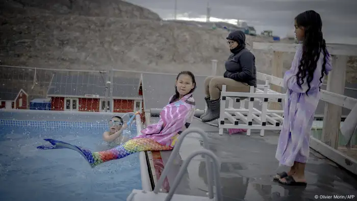 A girl sits at the edge of a pool with the tail of her mermaid costume, another child is in the water, a third in bathrobe and slippers watches. A woman sits next to her in fur boots and anorak.