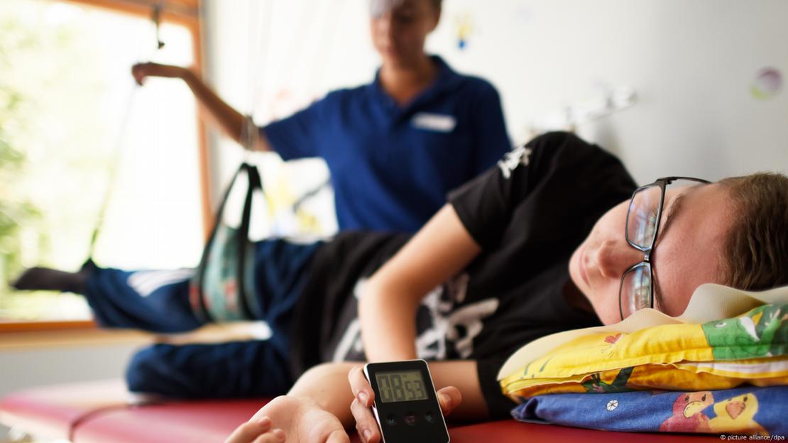 Criança deitada em leito de clínica sendo ajudada por enfermeira a fazer exercício