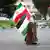 Ein muslimischer Mann läuft eine Straße entlang und trägt eine palästinensische und eine iranische Flagge 