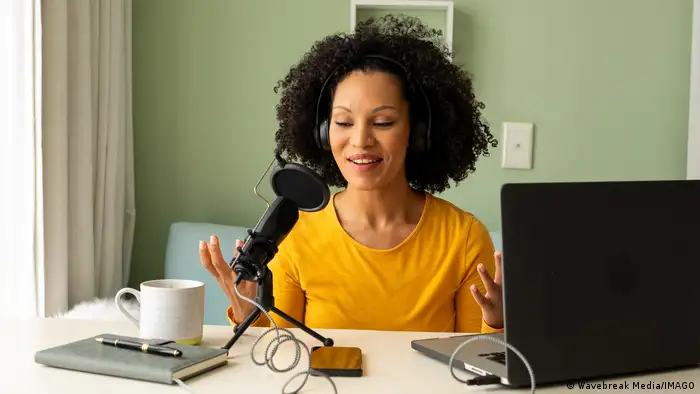 Aa woman sits at a desk with a laptop and a microphone in front of her