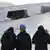 Drei Männer stehen im Schnee, dick vermummt in Jacken und Mützen bzw. einen Turban