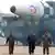 Kim Jong-un, de óculos escuros e jaqueta de couro preta, caminha entre dois miltares em frente a um veículo militar
