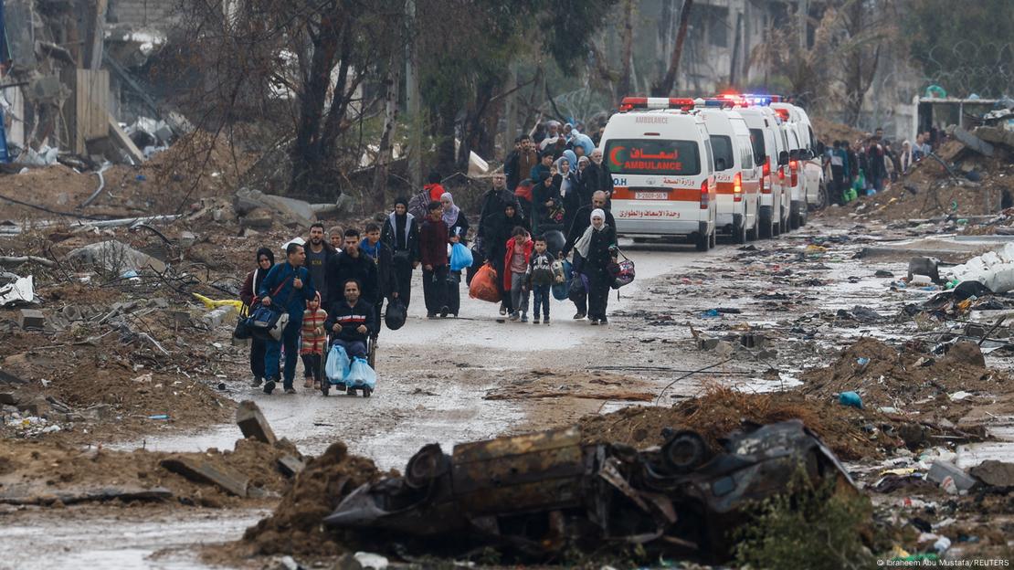 Pessoas com sacos de coisas caminham perto de ambulância em meia a uma área devastada.