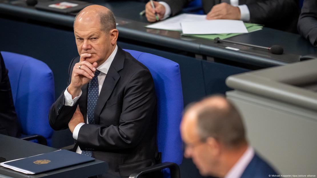 Bundeskanzler Olaf Scholz (SPD, l) sitzt im Bundestag auf einem der blau bezogenen Sessel der Regierungsbank und schaut zu Friedrich Merz, CDU-Bundesvorsitzender und Unionsfraktionsvorsitzender, der am Rednerpult steht. Merz Kopf ist im Vordergrund verschwommen zu sehen. Scholz hat die rechte Hand am Kinn. Vor ihm liegt eine blaue Mappe, die mit dem goldenen Bundesadler verziert ist. 