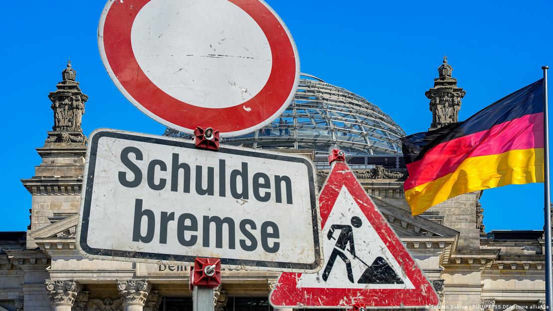 Verkehrsschilder Baustelle, Durchfahrt verboten, Wort "Schuldenbremse" vor dem Reichstagsgebäude und der deutschen Flagge
