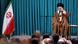  Ayatollah Ali Khamenei, in black turban and a brown robe, raises his left hand and smiles. He is sitting on the right of a podium with the Iranian flag on the left.