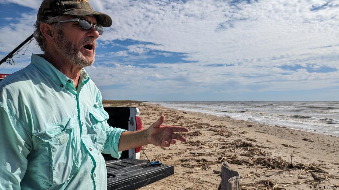 An older man stands on a beach beside his truck, wearing a ball cap and sunglasses
