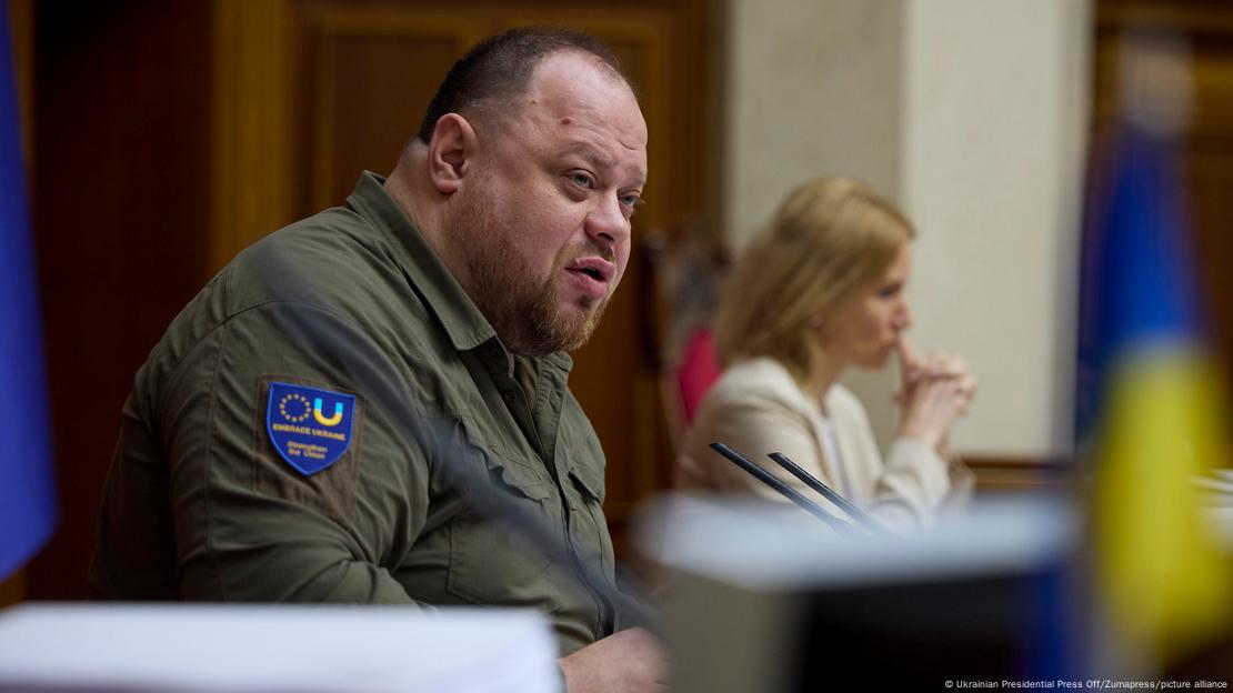 Ruslan Stefantschuk, seit Oktober 2021 Vorsitzender des ukrainischen Parlaments, sitzt an seinem Platz im Parlament und spricht in Mikrofone. Im Hintergrund ist eine Frau zu sehen.