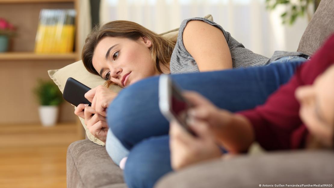 Duas mulheres jovens deitadas no sofá, cada uma olhando para o seu smartphone
