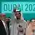 Vor dem Banner "Dubai 2023" stehen lachend zwei Männer und eine Frau vor Konferenzteilnehmenden, der Mann in der Mitte streckt Arm und Daumen hoch