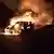 A silhueta de dois bombeiros é vista na escuridão contra as chamas de um carro ao fundo