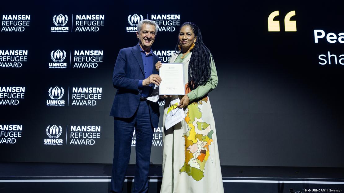 Mujer vestida de largo recibe premio de manos de hombre con traje.