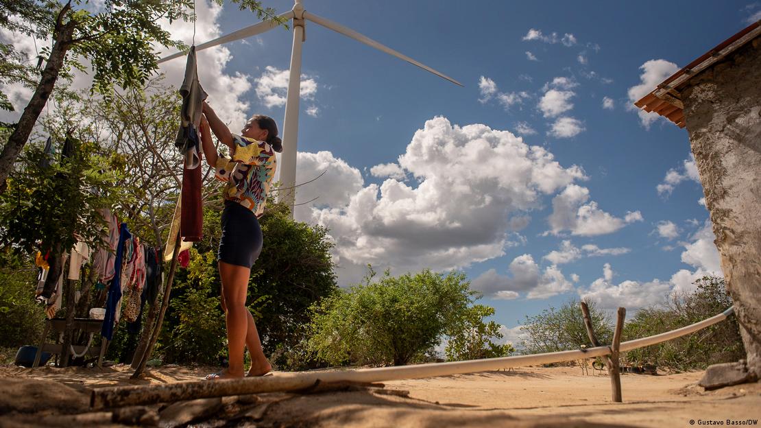 Maria do Socorro pendura roupas no quintal de sua casa. Ao fundo, turbina de geração de energia eólica.