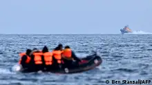 Manş Denizi'nde bir şişme bot içinde, üzerlerinde can kurtarıcı yelek olan insanlar, arka planda askeri bir gemi