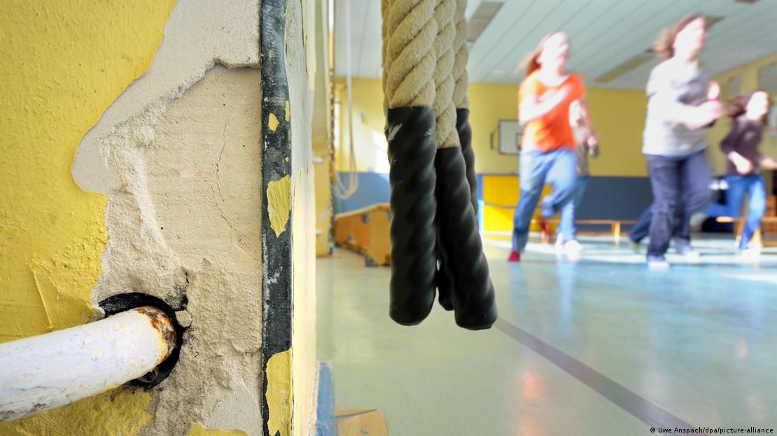 Aufgeplatzte Betonwand in einer Sporthalle während im Hintergrunde Schulsport läuft