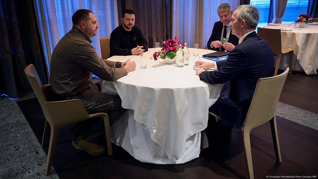 Zelenski sentado à mesa ao lado de três homens, um deles Jens Stoltenberg