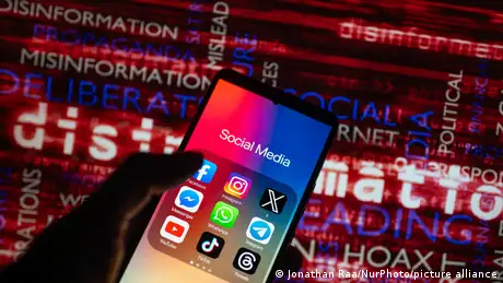 The logos of social media icons, such as Facebook, TikTok, WhatsApp, YouTube, X, Instagram, are displayed on a smartphone. A hand holds the phone in front of a screen showing words such as disinformation and misinformation.