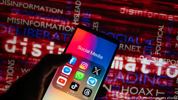 The logos of social media icons, such as Facebook, TikTok, WhatsApp, YouTube, X, Instagram, are displayed on a smartphone. A hand holds the phone in front of a screen showing words such as disinformation and misinformation.
