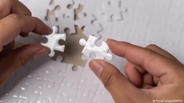 Two hands hold up two pieces of a completely white jigsaw puzzle with more puzzle pieces visible in the background