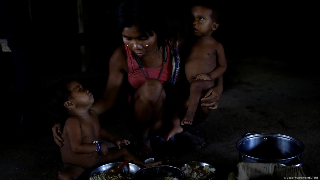 Mulher yanomami carrega criança no colo, enquanto abraça outro no chão