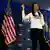 A candidata republicana Nikki Haley fala em microfone ao lado de duas bandeiras dos Estados Unidos.