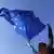 Pessoa segura com as duas mãos levantadas uma bandeira da UE com céu azul ao fundo