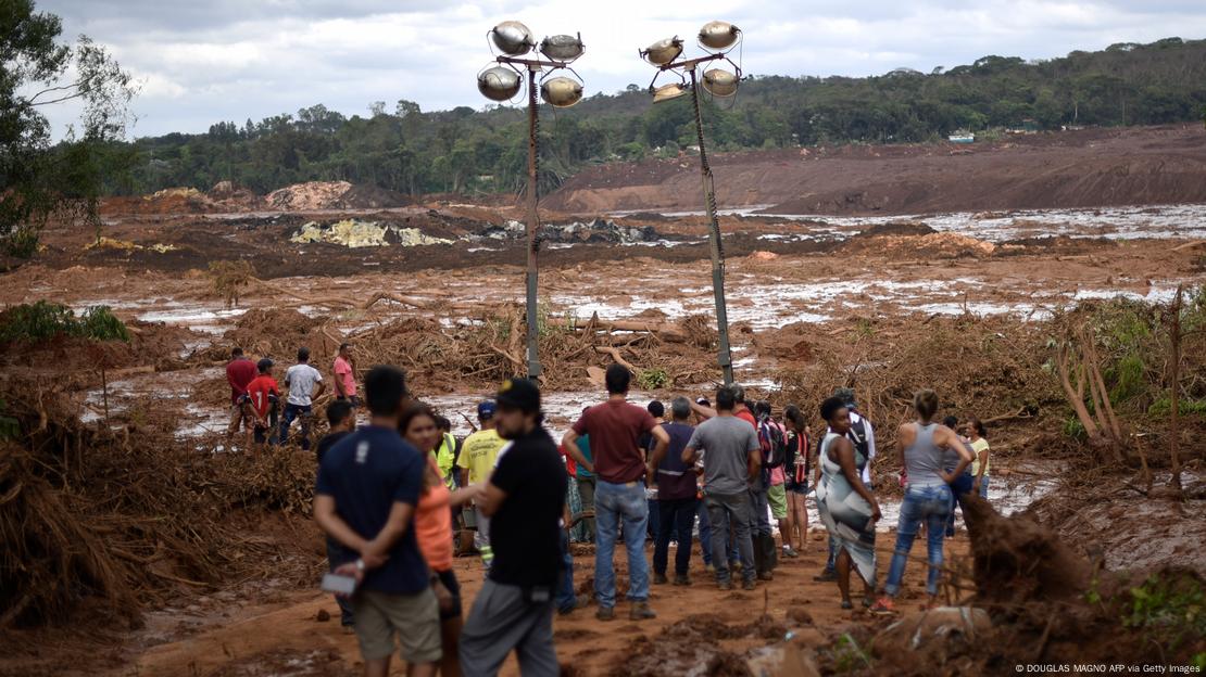 Foto de barragem colapsada em Brumadinho, com pessoas em primeiro plano olhando para a lama