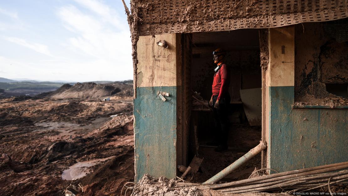 Imagem de bombeiro olhando para destruição causada no Córrego do Feijão, perto Brumadinho. Ele está dentro de uma casa manchada de marrom, do lado direito da imagem.